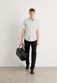 Cămașă verde deschis cu mânecă scurtă și un model subtil, pantaloni negri slim-fit și geantă de voiaj neagră, purtată cu pantofi negri pe un fundal neutru.
