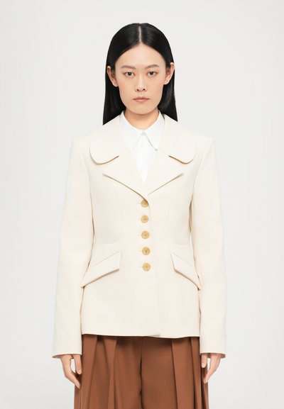Beige strukturierter Blazer mit breitem Kragen, fünf goldenen Knöpfen und zwei Seitentaschen, kombiniert mit einem weißen Hemd und braunen Hosen.