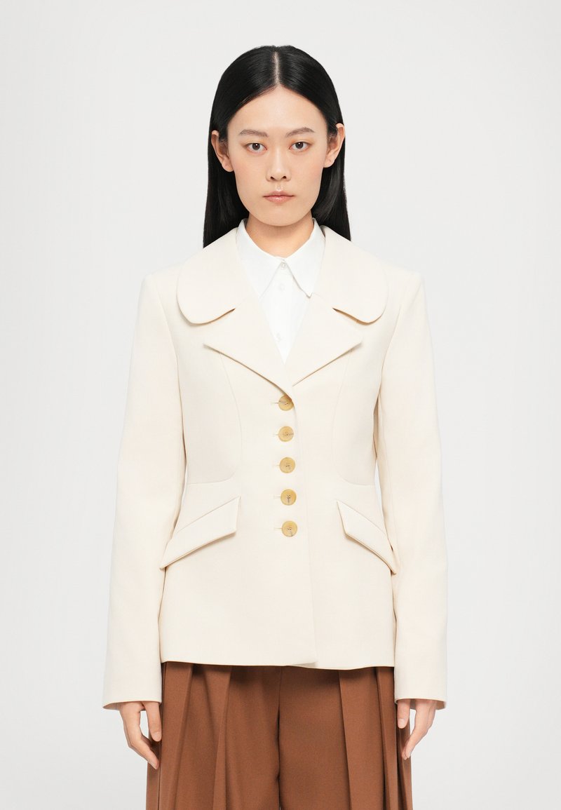 Blazer estructurado beige con un cuello ancho, cinco botones dorados y dos bolsillos laterales, acompañado de una camisa blanca y pantalones marrones.