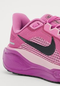 Ružičasta mrežasta atletska cipela s crnim Nike svušom; ima podstavljeni ovratnik, sistem vezanja i teksturiranu gumenu podlogu u nijansama ružičaste i crne.
