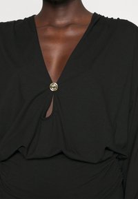 Black top with a v-neckline, featuring a twist detail and large gold button at the center. Soft, smooth fabric with a relaxed fit.
