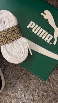 Bijele najlonske vezice za cipele, savijene i pričvršćene uzorkovanom Puma trakicom, naslonjene na zelenu Puma kutiju za cipele s bijelim logotipom.