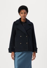 Marineblå dobbeltspent ullfrakk med store revers, gullfargede knapper og brede mansjetter, brukt over et satengskjørt. Glatt tekstur, strukturert design.