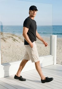 Homme avec casquette noire, t-shirt noir, short beige et chaussures noires marchant sur une terrasse en bois blanc près de la plage avec l'océan en arrière-plan.