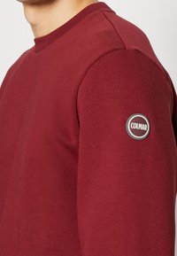 Felpa rossa con collo rotondo, tessuto testurizzato e una toppa circolare grigia sul maniche sinistro. Vestibilità sartoriale e aspetto liscia.