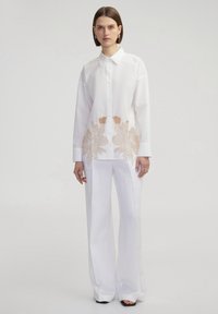 Camisa blanca de algodón con collar y mangas largas, que presenta intrincados bordados florales en tono beige en el dobladillo. Combinada con pantalones anchos blancos.