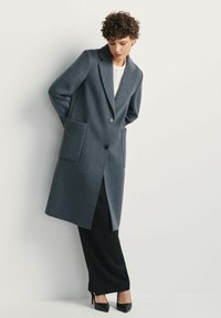 Kobieta z krótkimi kręconymi włosami, ubrana w długi szary płaszcz, biały top, czarne spodnie i czarne szpilki, stojąca przy białej ścianie.