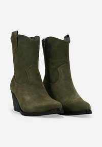 Par de botas de tobillo de ante verde oliva con cremalleras laterales, costuras decorativas, tiradores y tacones apilados en un fondo blanco.