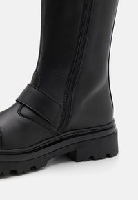 Botte mi-mollet en cuir noir avec fermeture éclair latérale, détail de sangle, semelle en caoutchouc robuste, surface texturée et talon plat.