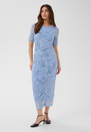 Vrouw draagt een lichtblauwe, gerimpelde, mid-lange jurk met korte mouwen en bloemenpatroon, gecombineerd met zwarte T-strap pumps, staand tegen een witte achtergrond.