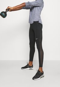 Černé sportovní legíny s síťovinovými panely, v kombinaci s modrým tričkem s dlouhým rukávem. Osoba drží černou činku. Černé tenisky s bílým akcentem.