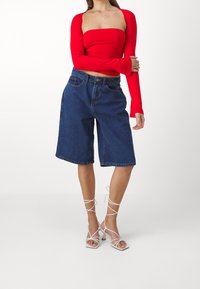 Červený dlouhý rukáv úzký top spolu s mid-blue denimovými šortkami. Model má na sobě bílé sandály na podpatku, což ukazuje na casualní, uvolněný outfit.
