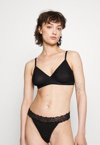 Bralette noire avec de fines bretelles réglables, présentant une forme triangulaire et une texture lisse. String noir avec des bordures en dentelle et des accents floraux.