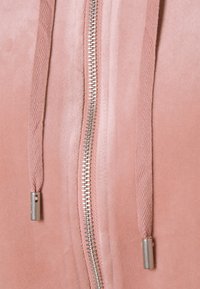 Felpa zip lucida di colore rosa chiaro, realizzata in tessuto morbido, con una cerniera metallizzata e coulisse coordinate con estremità texturizzate.