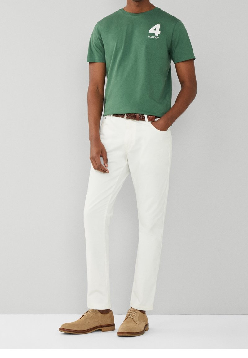 Camiseta de algodón verde con logo blanco "4", pantalones blancos, cinturón trenzado marrón y zapatos de ante beige. Pose de pie contra un fondo gris.