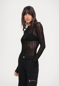 Body a maniche lunghe nero realizzato in tessuto trasparente con motivi ondulati. Abbinato a pantaloni cargo neri aderenti con zip laterali e tasche.