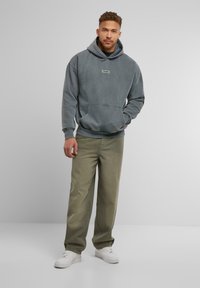 Grau Hoodie mit Kängurutasche und Schildpatch, kombiniert mit olivgrünen Hosen und weißen Sneakers. Weicher Stoff und lockere Passform.