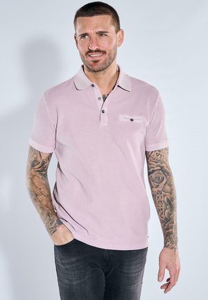 Man with short hair and tattoos on both arms wears light pink polo shirt and dark jeans, standing with one hand in pocket.