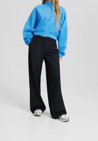 Persona con sudadera azul brillante con cremallera, pantalones anchos negros y zapatillas grises, de pie con las manos en los bolsillos sobre un fondo liso.