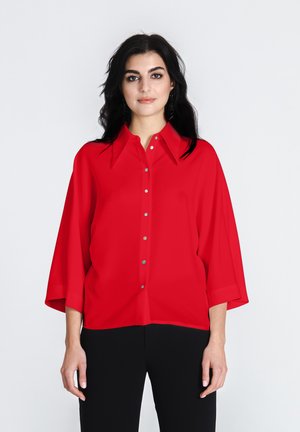 Rode overhemdd blouse met een kraag, korte mouwen en een relaxte pasvorm. Voorzien van metalen knopen en een gladde textuur. Gepaard met zwarte broeken.