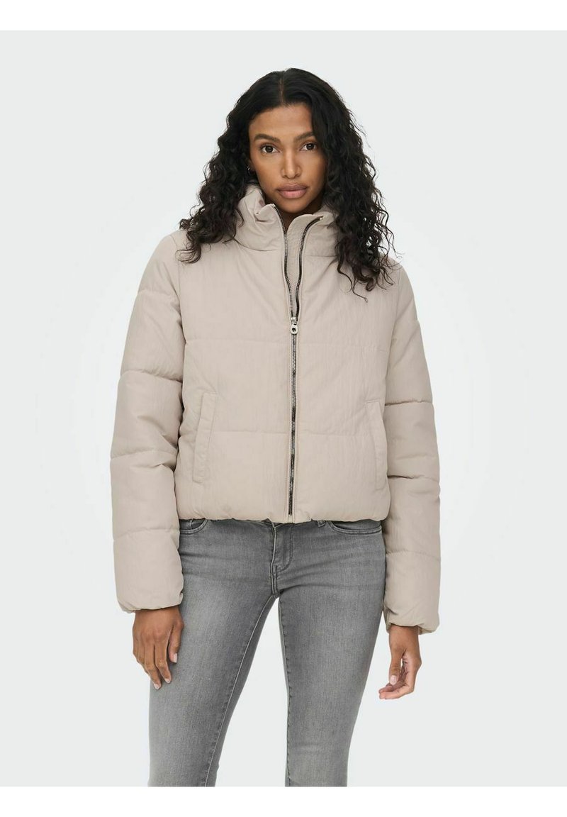 Giacca a vento in beige chiaro con colletto alto, zip frontale e design trapuntato. Abbinata a jeans grigi, mostra un look casual.