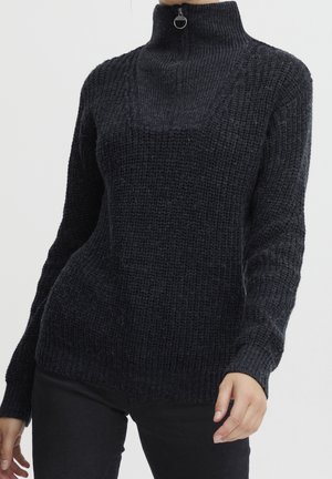 Femme portant un pull en maille texturée noir avec un col haut et un détail zippé, associé à un pantalon noir sur un fond uni.