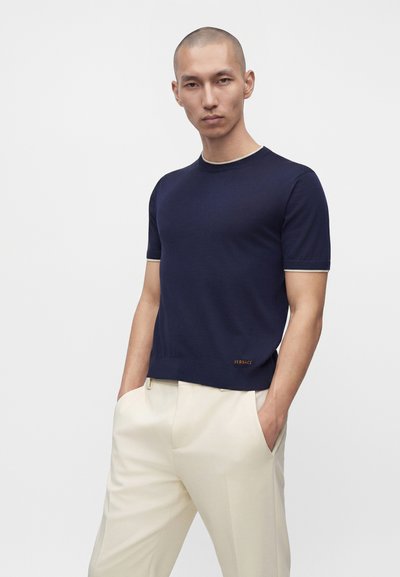 Homme au crâne rasé portant une chemise à manches courtes bleu marine et un pantalon crème, debout les mains dans les poches sur un fond uni.