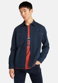 Navyblauwe button-up shirt van een zachte stof, met twee borstzakken en een kraag, gelaagd over een rode graphic t-shirt.