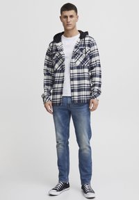 Camicia in flanella a quadri blu navy e bianca con cappuccio nero, abbinata a jeans blu e sneakers nere. Due tasche sul petto visibili.