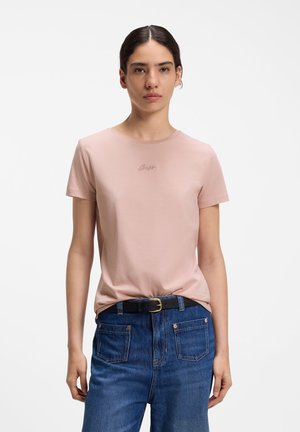 Kobieta o ciemnych włosach, ubrana w jasnoróżowy T-shirt z krótkim rękawem, wpuszczony w niebieskie dżinsy z czarnym paskiem, stojąca na białym tle.