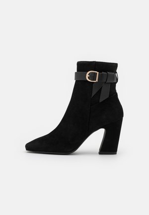 Bottines en suède noir avec un bout pointu, un talon bloc haut et une ceinture noire ornée d'une boucle dorée. Texture lisse dans l'ensemble.