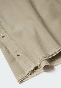 Tessuto di cotone beige con bordo sfilacciato, caratterizzato da una fila di bottoni visibili e cuciture bianche a contrasto lungo il lato.