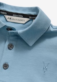 Ljusblå pikétröja av texturerat tyg. Har en vikbar krage, tre knappar och en diskret broderad getskallelogga.