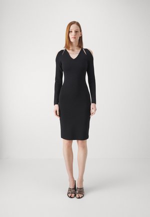 Black long-sleeve dress with a V-neckline and cut-out shoulders. Fitted shape and soft texture. Worn with black strappy heeled sandals.