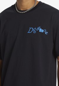 Zwarte katoenen t-shirt met een vervormd blauw logo "Dcshoe co" op de linkerkant van de borst. Korte mouwen, losse pasvorm en een gladde textuur.