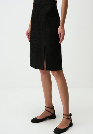 Black pencil skirt with textured fabric, knee-length, features a front slit, paired with black flat shoes and ankle straps.