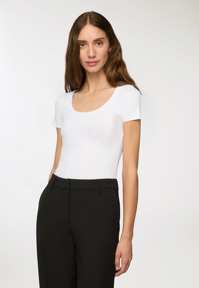 Top bianco a maniche corte a costine con scollo rotondo, abbinato a pantaloni neri su misura, su uno sfondo chiaro e semplice.
