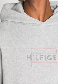 Felpa grigia realizzata in tessuto morbido con un grande logo "HILFIGE" incorniciato in rosso. Il design include una vestibilità comoda e un cappuccio con cordoncino.