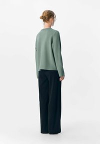 Femme aux cheveux blonds coiffés en chignon, portant un haut long à manches longues vert ample et un pantalon large noir, se tenant de dos devant un fond blanc.