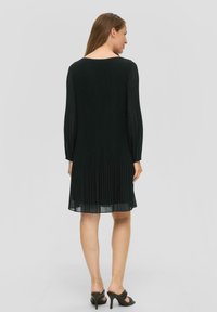 Schwarzes plissiertes Kleid mit langen Ärmeln und rundem Halsausschnitt. Der Stoff ist weich, mit einem dezenten Glanz und fließender Textur. Passt zu hohen Schuhen.