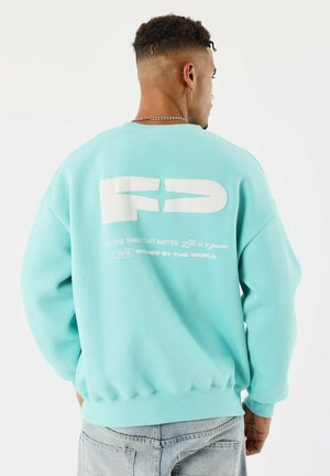 Uomo di spalle che indossa una felpa leggera blu con lettere bianche in grassetto "FP" e il testo "Fai le cose che contano" sul retro, jeans chiari e collana a catena.