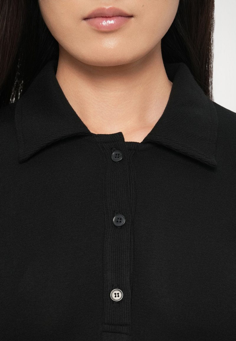 Zwart overhemd met kraag, voorzien van een geribbelde structuur, drie knopen aan de voorkant en een gladde halslijn. De stof oogt zacht en nauwsluitend.
