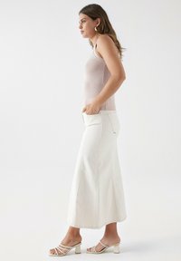 Una blusa sin mangas de color rosa claro combinada con una larga falda de mezclilla blanca que presenta bolsillos y un ligero acampanado, complementada con sandalias de tacón bloque blanco.