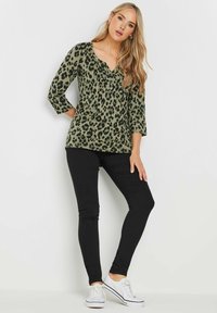 Grünes Leopardenmuster-Oberteil mit Dreiviertelärmeln und V-Ausschnitt, kombiniert mit schwarzen Skinny-Jeans und weißen Turnschuhen.