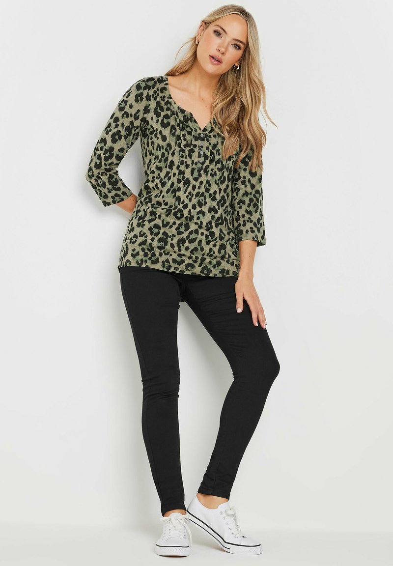Groen luipaardprint top met driekwart mouwen en V-hals, gecombineerd met zwarte skinny jeans en witte sneakers.