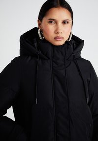 Veste matelassée noire avec un col montant, des cordons de serrage ajustables et deux poches avant. Texture lisse avec des détails en motif matelassé.