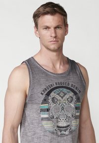 Siva brezrokavnik z osrednjim grafičnim motivom okrasne lobanje z geometrijskimi vzorci in turkiznimi detajli. Blago se zdi mehko.