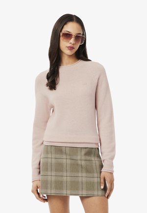 Maglione a maglia rosa chiaro con maniche lunghe e polsini a coste, abbinato a una mini gonna a quadri verdi. Modella con occhiali da sole traslucidi.