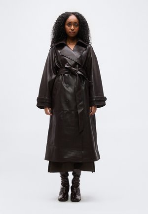 Γυναίκα με σγουρά μαλλιά, φοράει μακρύ καφέ σκούρο δερμάτινο παλτό τύπου τρανσέ σε ζώνη στη μέση, συνδυασμένο με κατάλληλα σκουρόχρωμα μπότες, στέκεται μπροστά σε λευκό φόντο.