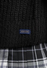 Schwarzer Strickpullover mit gerippter Textur und blauer Etikette. Kombiniert mit einem karierten Rock, der schwarz-weiß-graue Muster aufweist.